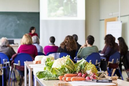 Personen in einem Seminar zur gesunden Ernährung sitzen mit dem Rücken zur Kamera, im Vordergrund ist Gemüse auf einem Tisch zu sehen.