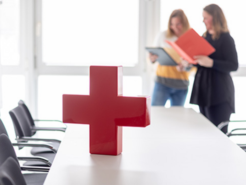 Das Rote Kreuz steht auf einem Tisch und zwei Frauen lesen im Hintergrund. 